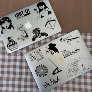 May include: Two silver laptops with black and white stickers featuring characters and text from the anime series Neon Genesis Evangelion. The stickers include the text "UNIT-01", "PROJECT EVA", "NEO TOKYO", "3RD IMPACT", "WARNING", "NE", "R", "NEON GENESIS", "EVANGELION", and "エヴァンゲリオン".