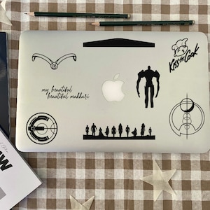 May include: A silver laptop with various black stickers, including a silhouette of a robot, a stylized apple logo, a circle with a crescent moon, a target with a letter C in the center, a group of people walking, and the text "Kiss the Cook" with a chef's hat. The text "my beautiful beautiful makkaari" is also written on the laptop.