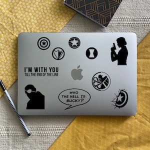 Puede incluir: Un ordenador portátil plateado cubierto de pegatinas negras con temática de superhéroes. Las pegatinas incluyen emblemas de escudo, una silueta de un personaje con una pistola y texto que dice "I'M WITH YOU TILL THE END OF THE LINE" y "WHO THE HELL IS BUCKY?"