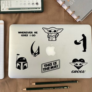 Könnte beinhalten: Ein silberner Laptop mit mehreren schwarzen Vinyl-Aufklebern mit Figuren und Sätzen aus dem Star Wars-Franchise. Die Aufkleber zeigen einen Charakter aus der Serie The Mandalorian, ein Zitat aus der Serie und die Worte "This is the way".