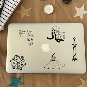 Puede incluir: Un portátil blanco con varias pegatinas en blanco y negro. Las pegatinas incluyen un personaje de dibujos animados, una calavera y una espada, una mujer con un vestido y una pegatina circular que dice "Juntos en París". El portátil también tiene texto escrito a mano que dice "Dimitri Anya" y marcas de conteo.
