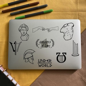 May include: A silver laptop with black and white stickers. The stickers feature a trident, a helmet, a lyre, a column, a hand reaching out, a bust of a woman, a torso, and the text "goddess of love" and "under world".