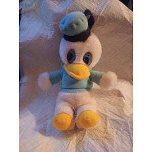 Könnte beinhalten: Ein Plüschtier von Donald Duck. Das Spielzeug hat einen weißen Körper, ein blaues Hemd und einen Hut sowie einen gelben Schnabel und Füße. Der Hut hat ein schwarzes Band. Das Spielzeug hat große blaue Augen und sitzt auf einer weißen Oberfläche.