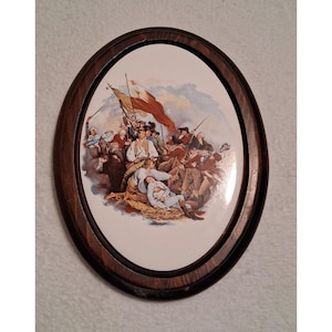 May include: Oval-shaped decorative plaque with a historical painting depicting a battle scene, framed in dark wood. The painting features soldiers, a flag, and the text "Bunker's Hill John Trumbull". The artwork is set against a white background.