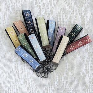 Puede incluir: Una colección de llaveros de tela tipo pulsera en varios colores, incluyendo amarillo, melocotón, azul oscuro, verde, azul claro, lavanda y rojo. Cada correa presenta un patrón floral blanco repetitivo y está unida a un mosquetón de metal oscuro.