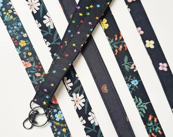 May include: A collection of fabric lanyards featuring various patterns on a black background, including white flowers, colorful small stars, and tiny rainbows. Each strap is finished with a silver-toned metal swivel clasp and a split ring.