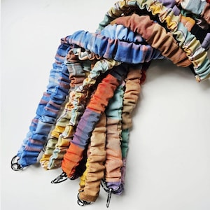 May include: A selection of colourful, fabric-covered cords with metal rings at the ends. The cords are in shades of blue, orange, yellow, and brown. The fabric appears to be gathered or ruched, creating a textured effect. The cords are arranged in a bundle.