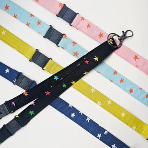 Peut inclure: Une collection de cordons en tissu avec des motifs d'étoiles dans diverses couleurs, notamment noir, jaune, bleu clair, bleu foncé et rose. Chaque cordon comprend une boucle en plastique noir et un mousqueton en métal pour attacher des objets.