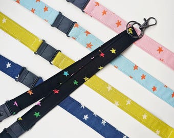May include: A collection of fabric lanyards featuring star patterns in various colors including black, yellow, light blue, dark blue, and pink. Each lanyard includes a black plastic buckle and a metal lobster clasp for attaching items securely.