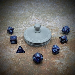 Könnte beinhalten: Ein Satz von sieben blauen und schwarzen Würfeln mit weißen Zahlen, angeordnet in einem Kreis um eine graue, dekorative Box mit einem Knopf oben.