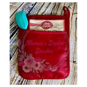 May include: Red oven mitt with a floral design and the text "Mumsi's Digital Doodles". A blue spatula and a Betty Crocker box are in the pocket of the mitt.