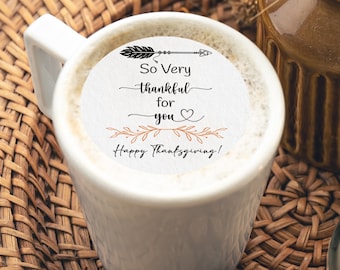 Op de afbeelding: Een witte mok met de tekst "So Very thankful for you Happy Thanksgiving!" staat op een geweven dienblad naast een brandende kaars in een glazen pot en een kleine bruine vaas met gedroogde planten. Dennenkegels zijn verspreid.
