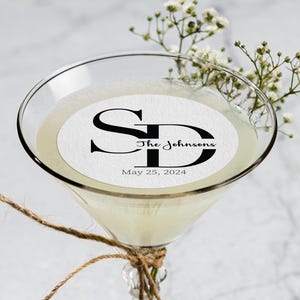 May include: A martini glass filled with a pale yellow liquid, featuring a custom edible topper with the initials "SD", "The Johnsons", and the date "May 25, 2024". The glass is adorned with twine and sprigs of white flowers.