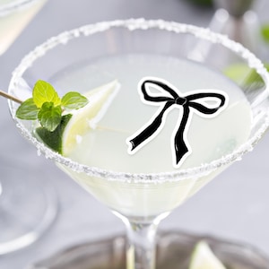 May include: A clear glass margarita glass with a sugared rim, filled with a pale yellow liquid. A lime wedge and a sprig of mint are in the glass. A black bow edible topper is on top of the drink. The glass is on a silver tray with lime wedges and mint leaves.