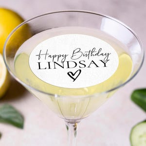 May include: A martini glass filled with a pale yellow liquid, topped with a white edible topper that reads "Happy Birthday LINDSAY" with a heart. The image includes lemons, cucumber slices, mint leaves, and a whole cucumber.