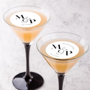 May include: Two martini glasses with black stems and clear bowls filled with a light orange liquid. Each glass has a white circular edible topper with the initials "M & P" and the date "08.14.2026". The Edible Design Co. logo is in the bottom right.