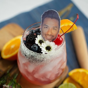 Puede incluir: Un cóctel rosa con hielo, adornado con una cereza, una rodaja de naranja, flores blancas y arándanos. Una foto de una persona está encima de la bebida.