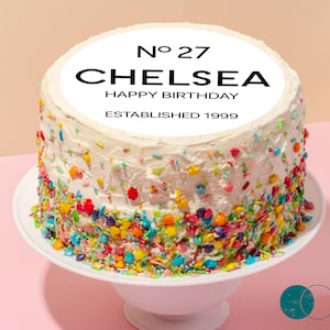 May include: A white frosted birthday cake with colorful sprinkles, sitting on a white cake stand. The cake has a circular topper with the text "No 27 CHELSEA HAPPY BIRTHDAY ESTABLISHED 1999". The cake is surrounded by gift boxes and a glass with red and white straws.