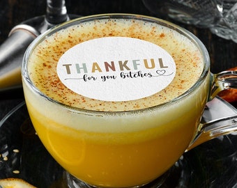 Op de afbeelding: Een heldere glazen kop met gele drank en een witte schuimlaag, op een schotel. De drank heeft een ronde eetbare topper met de woorden "THANKFUL for you bitches". Gegarneerd met sinaasappelschijfjes en munt.
