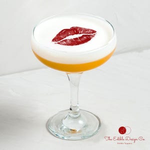 Puede incluir: Un c&oacute;ctel en una copa con una espuma blanca y una huella de labios rojos. La bebida tiene una base amarillo-naranja y un tallo de cristal transparente. El logotipo de Edible Design Co. est&aacute; en la esquina inferior derecha.