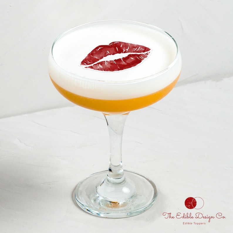 Puede incluir: Un c&oacute;ctel en una copa con una espuma blanca y una huella de labios rojos. La bebida tiene una base amarillo-naranja y un tallo de cristal transparente. El logotipo de Edible Design Co. est&aacute; en la esquina inferior derecha.