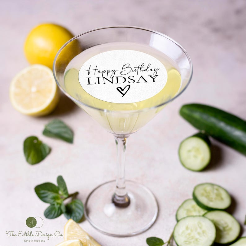May include: A martini glass filled with a pale yellow liquid, topped with a white edible topper that reads "Happy Birthday LINDSAY" with a heart. The image includes lemons, cucumber slices, mint leaves, and a whole cucumber.