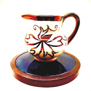 May include: A small, decorative ceramic pitcher with a handle and spout, featuring a white base with a floral design in red and black. The pitcher sits on a round, wooden-framed base with a dark blue center. The handle and rim are brown.