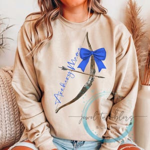 May include: A tan sweatshirt with a blue bow and a drawing of a bow and arrow with the text "Archery Mama" on it.