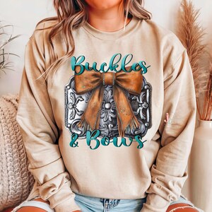 May include: A tan sweatshirt with a graphic design of a silver buckle with a brown bow and the text "Buckles & Bows" in teal blue.