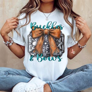 May include: White t-shirt with a graphic of a silver belt buckle with a brown bow and the text "Buckles & Bows".