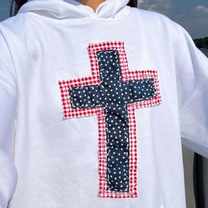 以下が含まれることがあります： 白いフード付きスウェットシャツ、十字のデザイン。 十字は白い星が付いた濃い青色の生地でできており、赤と白のギンガム生地で縁取られています。 スウェットシャツにはフロントポケットとドローストリングフードが付いています。