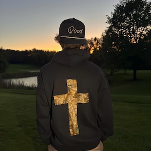Pode incluir: Moletom preto com uma grande cruz de camuflagem nas costas. Um boné preto com a palavra "Good" bordada em branco é usado. A imagem é tirada ao ar livre ao anoitecer, com um campo de golfe e árvores ao fundo.