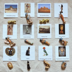 May include: A collection of twelve educational cards featuring images of Ancient Egyptian landmarks, deities, and artefacts, accompanied by twelve corresponding miniature figurines including a scarab, cat, falcon, and various human-like figures.
