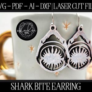 May include: Silver-colored shark bite earrings with detailed teeth and jaw design. The earrings are displayed against a light-colored mug with a gold handle. The image includes the text "SHARK BITE EARRING".