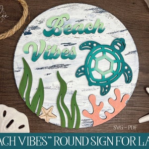 Op de afbeelding: Een rond houten bord met de tekst "Beach Vibes" in een groene gradiënt. Het bord heeft een groene en blauwe gradiënt schildpad, koraal en zeewier. Het bord is perfect voor een strandthema huisdecoratie.