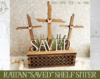 May include: A wooden shelf sitter with the word "SAVED" in large letters, surrounded by three crosses with doves on top. The crosses are set against a background of green leaves. The shelf sitter is made of a rattan-like material.