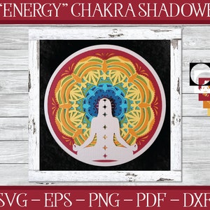 Op de afbeelding: Een 3D-papiercraft shadowbox met een mandala-ontwerp met een mediterend figuur in het midden. De mandala is gemaakt van zeven kleuren, waaronder rood, oranje, geel, groen, blauw, indigo en violet. De shadowbox is ingelijst in wit hout.