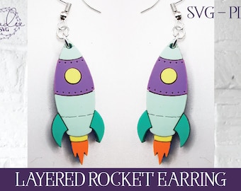 May include: A pair of layered rocket earrings. Each earring features a rocket design with a purple top, yellow circle, light blue body, teal fins, and orange flames. The earrings are suspended from silver-colored hooks. The text "SVG - PDF" is visible.
