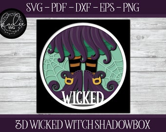 Könnte beinhalten: Eine 3D-Schattenbox mit Hexenbeinen und -füßen, die schwarze Stiefel mit goldenen Schnallen tragen. Der Hintergrund ist ein grünes und violettes Wirbelmuster mit einem Spinnennetzdesign. Der Text "WICKED" ist in Weiß auf der Vorderseite der Box gedruckt.
