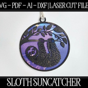 May include: A purple and blue sloth suncatcher with a black silhouette of a sloth hanging from a branch. The suncatcher is round and has a white string attached to the top.