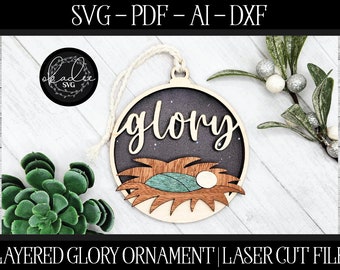 May include: A layered wooden Christmas ornament featuring the word "glory" in a cursive font. The ornament depicts a manger scene with a baby Jesus, a swaddling cloth, and hay. The ornament is designed for laser cutting.