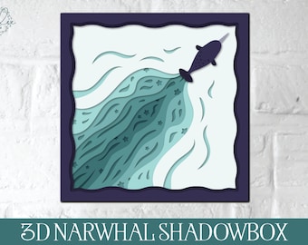 May include: A 3D papercraft shadowbox featuring a narwhal with a blue and green layered design. The narwhal is depicted swimming through a wave of water with stars.