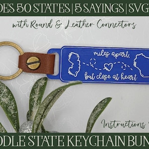 Puede incluir: Un llavero azul y blanco con una correa de cuero y un anillo para llaves. El llavero presenta un contorno de estado y el texto "miles apart, but close at heart".