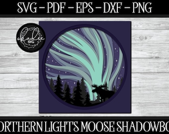 May include: A digital design for a shadowbox featuring a moose standing in front of a stylized depiction of the Northern Lights. The design is in shades of blue, green, and purple.