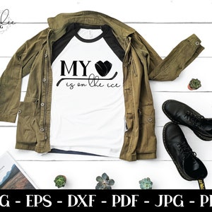 Pu&ograve; includere: Una maglietta bianca con una grafica di bastone da hockey nero e rosso e il testo "MY &hearts; is on the ice". La maglietta &egrave; su una superficie di legno con una giacca verde e stivali neri.