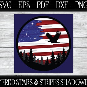 May include: A layered design depicting the American flag with stars, stripes, and an eagle in flight, framed in a circle. A forest silhouette is at the base. The text "LAYERED STARS & STRIPES SHADOWBOX" is at the bottom of the image.