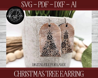 May include: A pair of wooden Christmas tree earrings with a distressed white paint finish. The earrings are laser cut with a detailed design of a Christmas tree with snowflakes and swirls. The earrings are hanging from silver hooks.