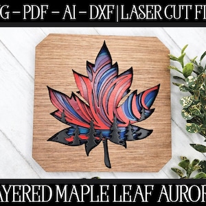 Puede incluir: Una hoja de arce de madera en capas con un diseño de aurora rojo, azul y negro. La hoja está sobre un fondo de madera.