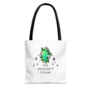 May include: White tote bag with black handles. The bag features a green gem design with white flowers and the text "let yourself bloom." This tote bag is made of a durable material and is suitable for carrying books, groceries, or personal items.