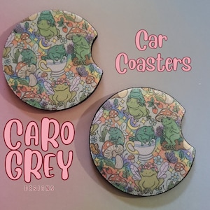 May include: Two round car coasters with a whimsical design featuring frogs, mushrooms, flowers, and stars on a light blue background. The text "Car Coasters" is written in pink letters on a white background.